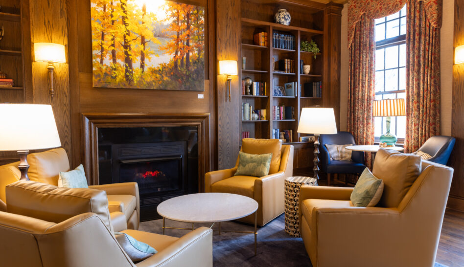 Elegant lounge with wood-paneled walls, bookshelves, armchairs, lamps, a fireplace, and a large autumn landscape painting.